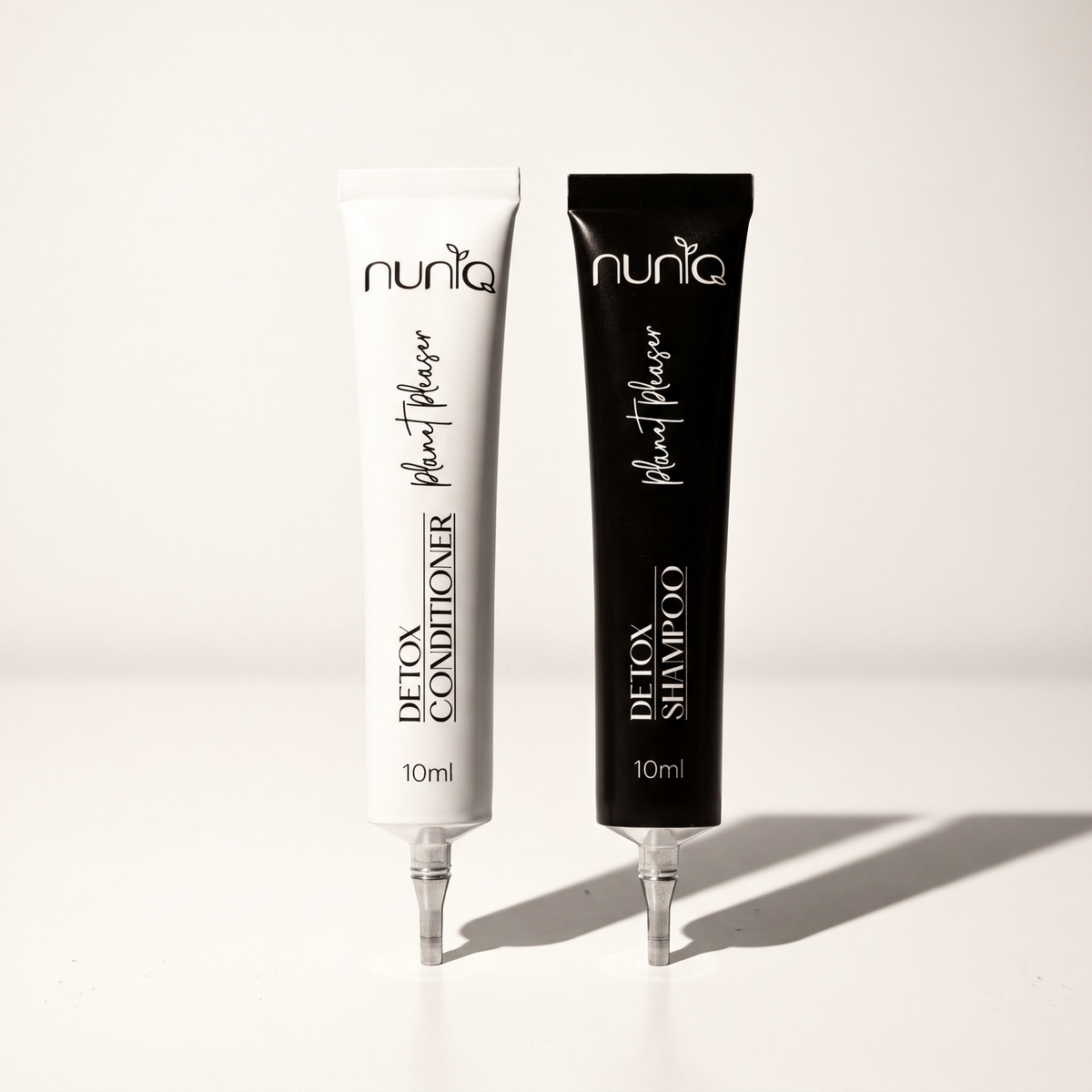 Two 10ml squeeze tubes of Nuniq Detox Shampoo and Detox Conditioner standing upright on a white surface with soft shadows. The shampoo tube is black and the conditioner tube is white, both with black and white text.