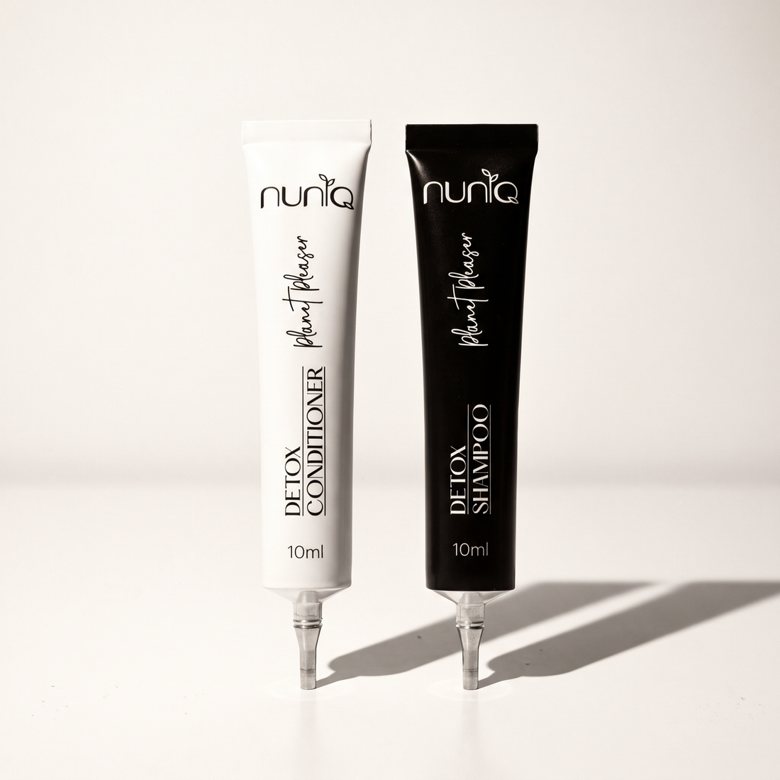 Two 10ml squeeze tubes of Nuniq Detox Shampoo and Detox Conditioner standing upright on a white surface with soft shadows. The shampoo tube is black and the conditioner tube is white, both with black and white text.