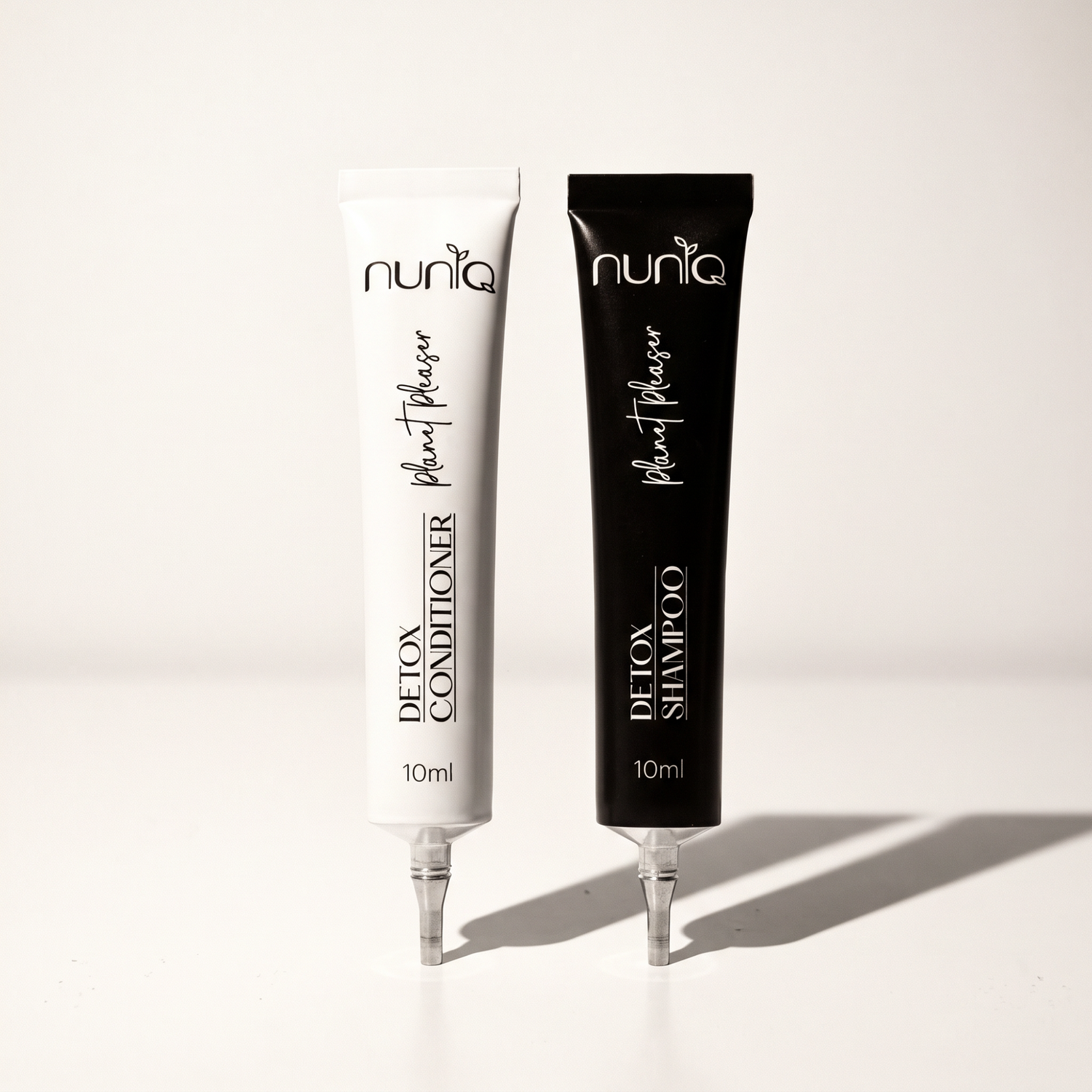 Two 10ml squeeze tubes of Nuniq Detox Shampoo and Detox Conditioner standing upright on a white surface with soft shadows. The shampoo tube is black and the conditioner tube is white, both with black and white text.