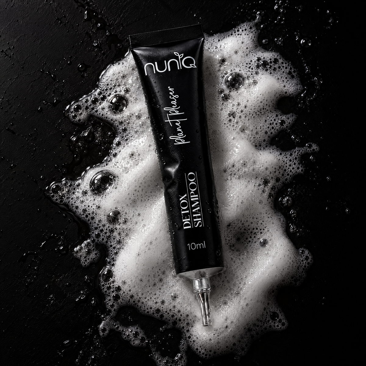 A black squeeze tube of Nuniq Plant Based Detox Shampoo lies diagonally on a glossy black surface surrounded by white foam bubbles.